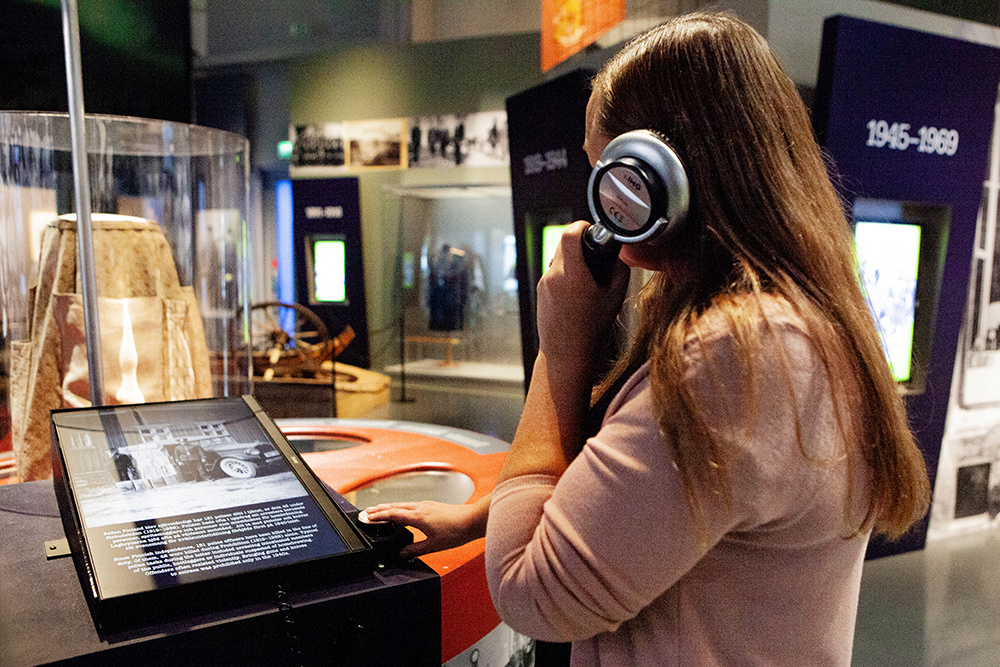 En person står med ryggen mot kameran och håller i hörluren. I bakgrunden syns museets utställningsutrymme.