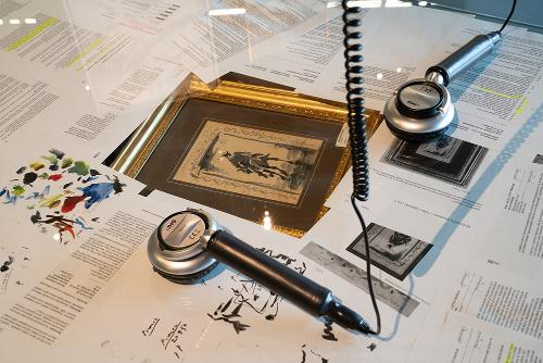 Written records and other pre-trial investigation materials spread on a table, with a framed painting in the middle. On top, two museum headsets, which museum visitors can use to listen to information about the exhibition.