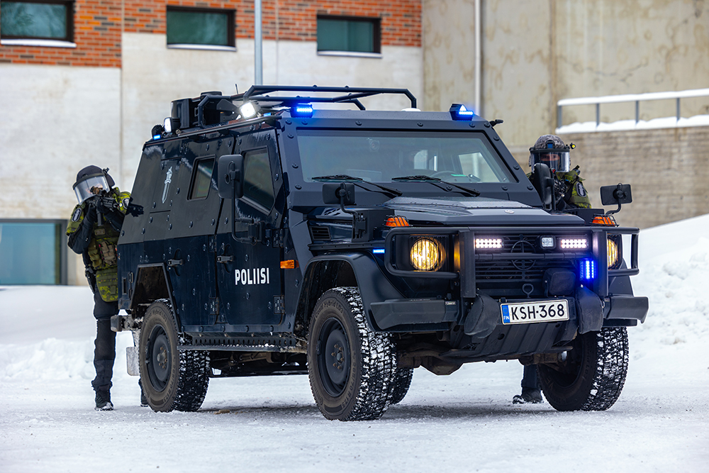 A dark armored off-roader type car stopped in front of a block of flats. ‘POLIISI’ is written on the side of the car. Police officers look out from behind the car, one on each side.