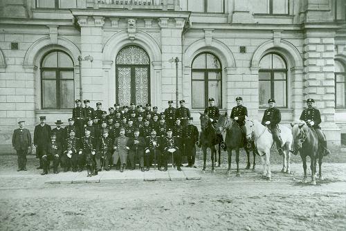 A black-and-white photograph of around twenty members of the Police Department staff posing for the camera in front of an old stone building. Some members of the group are sitting down and others are standing, while four persons are mounted on a horse. Most of the persons pictured are wearing a police uniform.