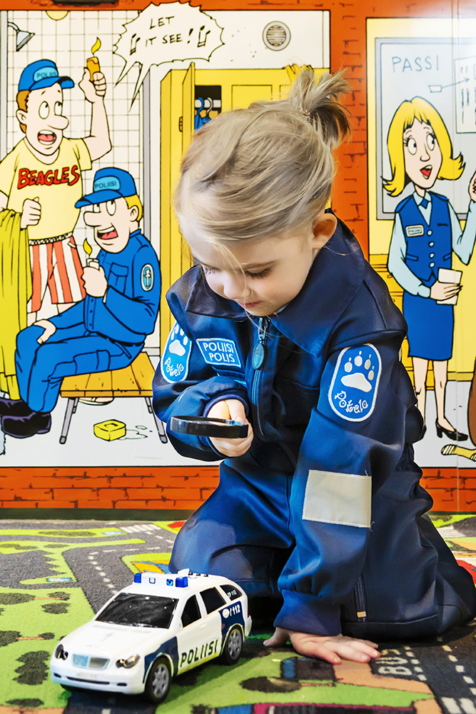 A child wearing police overalls is playing in the Police Museum’s children’s section, holding a magnifying glass and looking at a miniature police car. The child is sitting on a traffic play mat, and a wall featuring police-themed comics is seen in the background.
