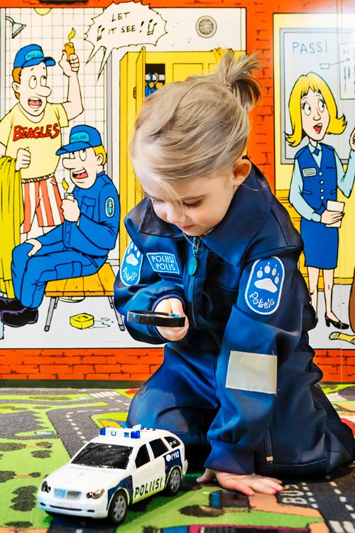 A child wearing police overalls is playing in the Police Museum’s children’s section, holding a magnifying glass and looking at a miniature police car. The child is sitting on a traffic play mat, and a wall featuring police-themed comics is seen in the background.