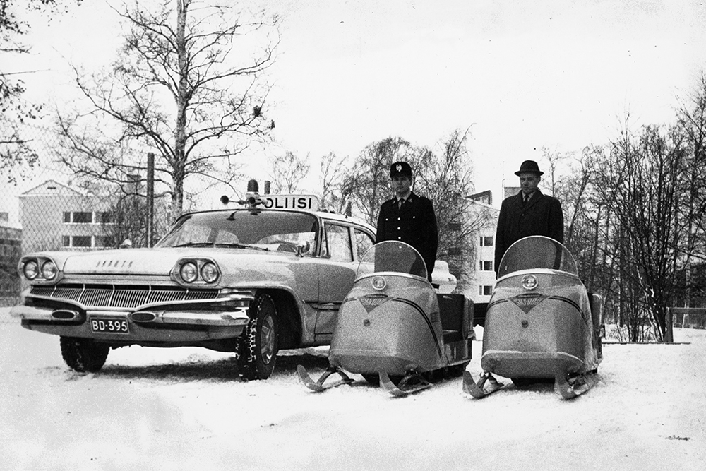 An old light-colored police car next to two old snowmobiles in winter landscape. Two persons pose on top of the snowmobiles.