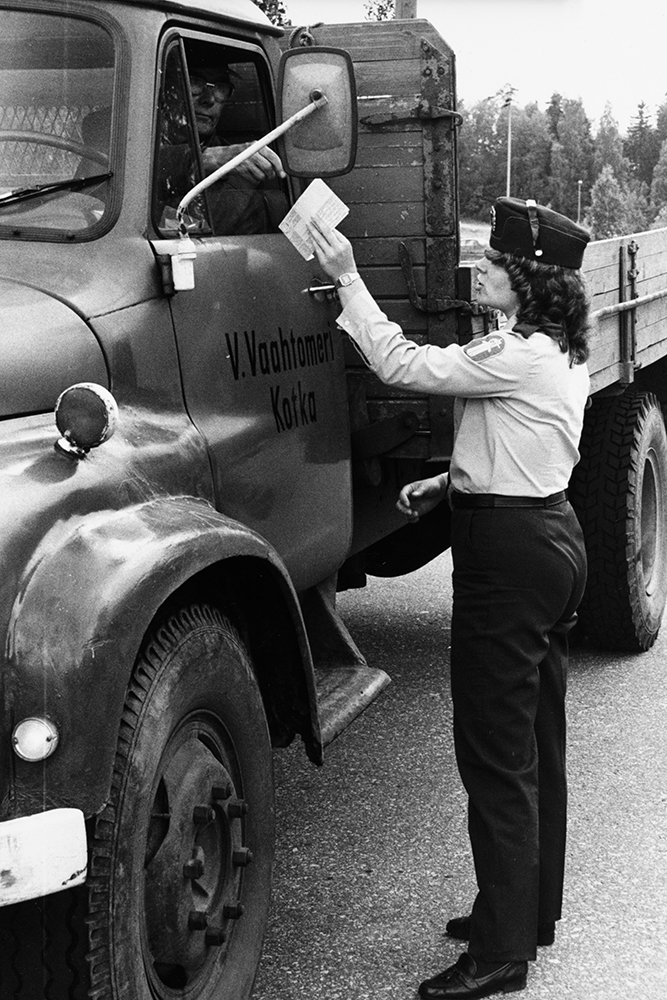 A black-and-white photograph of a female police officer in uniform checking the paperwork of a lorry driver.