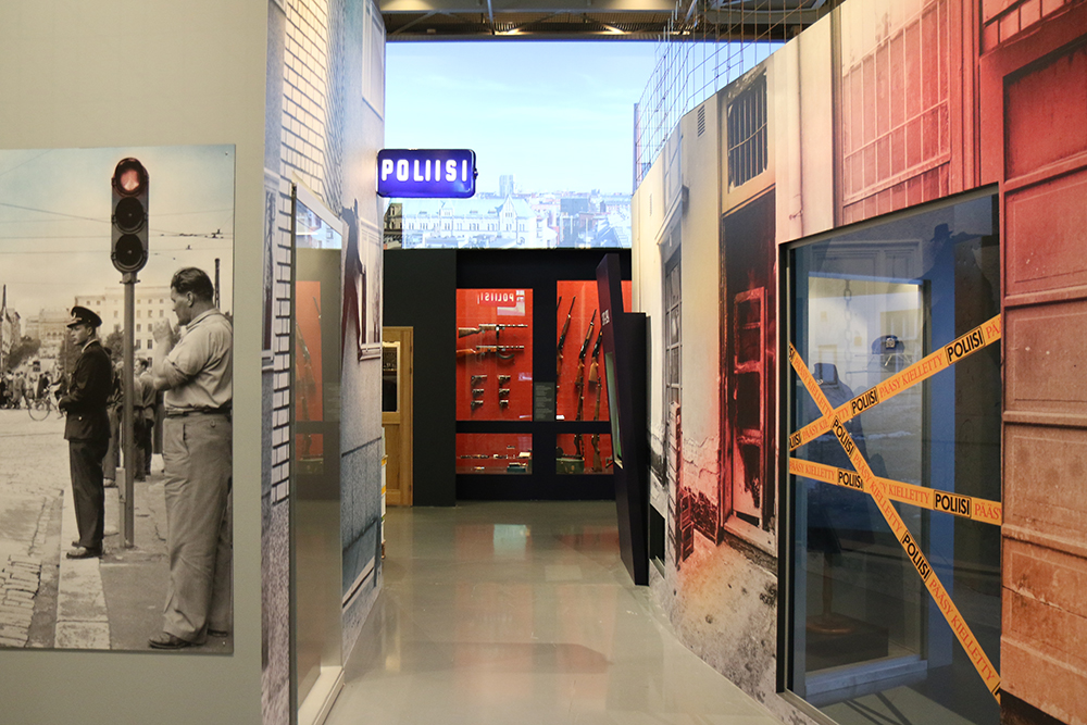 A view of the Museum’s exhibition. The picture shows, among others, the ‘Poliisi’ (the police) sign on the wall and some police tape “Police – do not cross”, and weapons on the wall in the background.