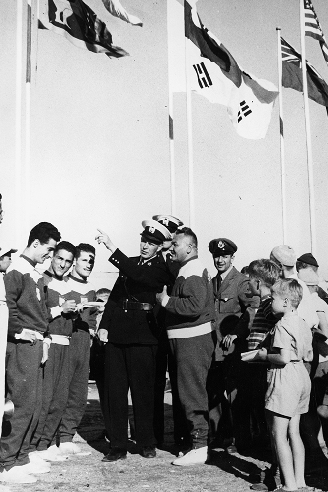 Three uniformed police officers are guiding Olympic athletes. The picture also shows children admiring the athletes. In the background, there are flagpoles with flags of various countries.