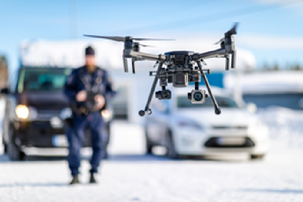 At the front, an unmanned aircraft of the police (UAS), a drone, is seen hovering in the air. A uniformed police officer controlling the UAS, and cars can be seen in the background.