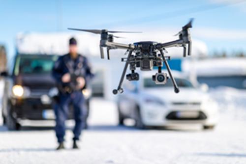 I förgrunden ser man polisens obemannade luftfartyg (UAS), det vill säga drönare, uppe i luften. I bakgrunden syns bilar och en polis i uniform som styr drönaren.