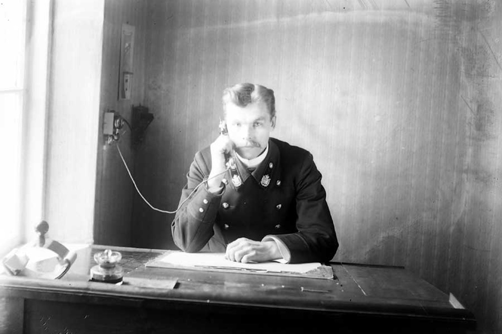 A black-and-white photograph of a police officer in uniform at a desk, making a telephone call.