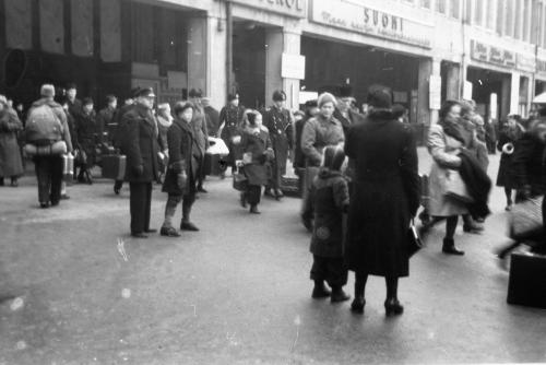 A black-and-white photograph of police officers patrolling at the Helsinki railway station, and adults and children with suitcases, standing and walking around.