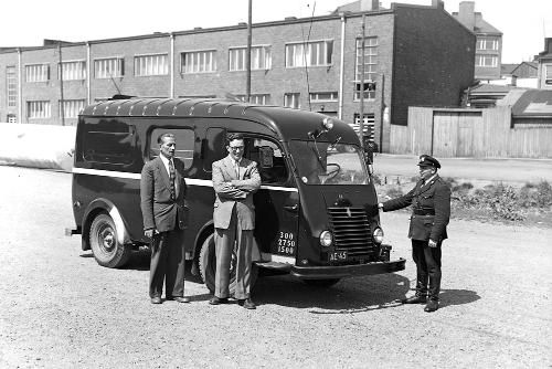 Mustavalkokuva, jossa kolme miestä seisoo pakettiauton edessä. Yhdellä miehistä on poliisin virkapuku ja kahdella siviiliasu. Taustalla näkyy tiilirakennus.