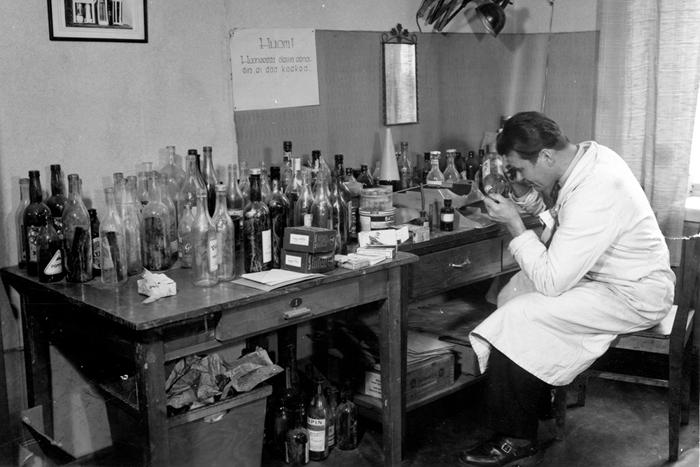 A black-and-white photograph of a man, wearing a lab coat, sitting at a desk, closely investigating a glass bottle. Dozens of different glass bottles stand next to him on the table.