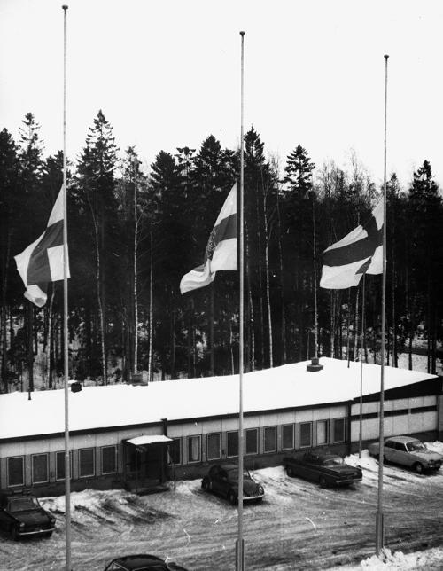 Black-and-white photograph with several flags flying at half-mast in front of a building in winter. Several cars stand parked in front of the building. There is forest in the background.