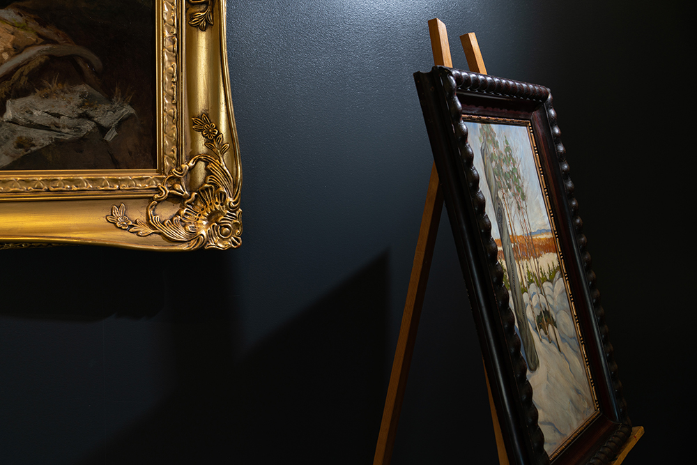 Two paintings in the Museum’s exhibition. One is hanging on the wall, and only the frame in gold and a small part of the dark artwork is seen. The other one is a landscape on an easel.