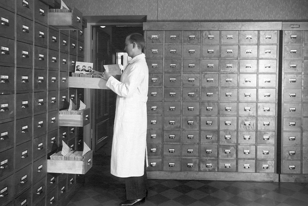 A black-and-white photograph of a man, wearing a lab coat, standing in an archive room. The walls of the room are stacked high with archival drawers. The man is examining a paper he has taken out of a drawer.