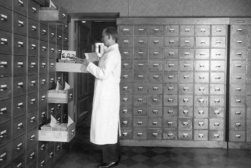 A black-and-white photograph of a man, wearing a lab coat, standing in an archive room. The walls of the room are stacked high with archival drawers. The man is examining a paper he has taken out of a drawer.