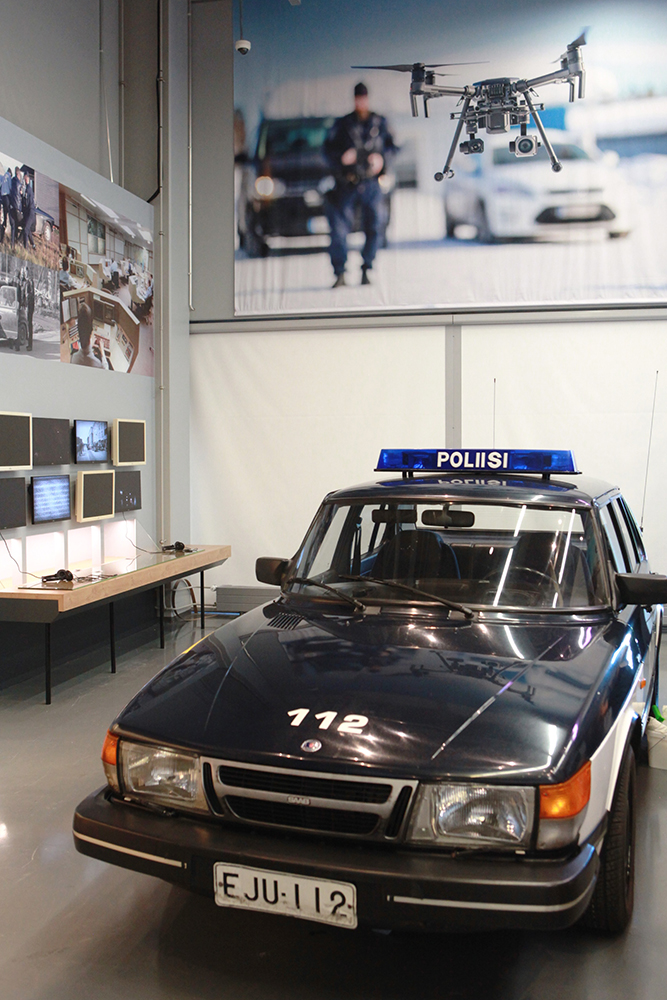 Inside the museum there is a dark blue Saab police car with a police sign on the roof. Computer screens and photos appear in the background.