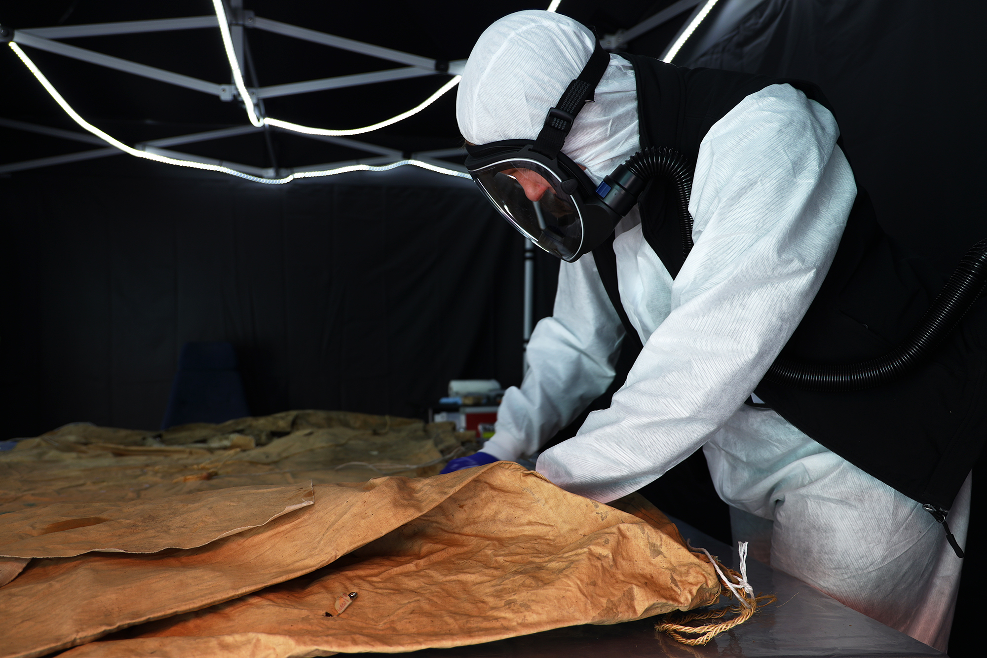A person dressed in a white protective suit and blue gloves examines the brown tent cloth on the table. The person has a respirator on their face.