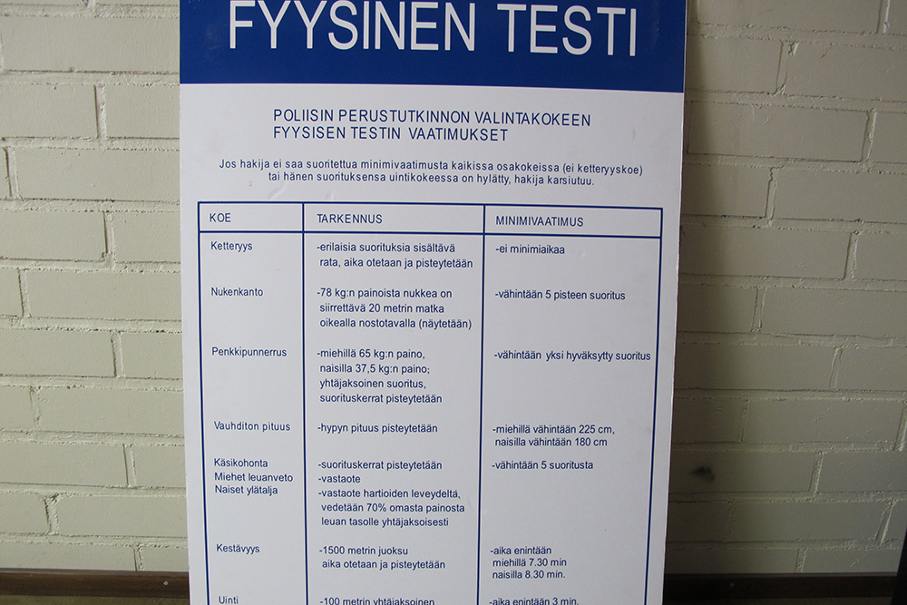 En färgbild av en skylt med instruktioner och krav för det fysiska testet inför polisens urvalsprov. Skylten innehåller specifikationer och minimikrav för smidighet, dockbärande, bänkpress, stående längdhopp, räckhäv/latsdrag för kvinnor, uthållighet och simning.