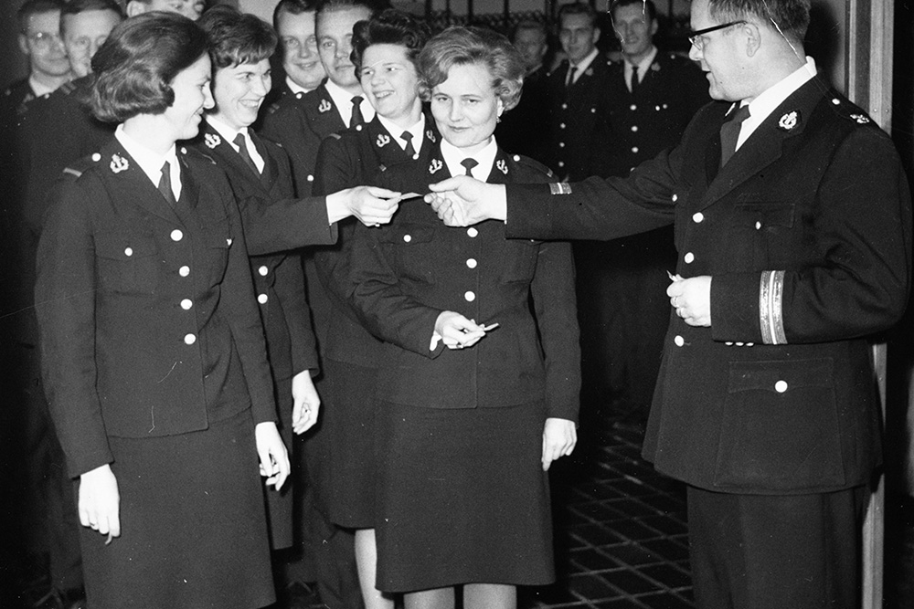 Svartvitt foto av en interiör, med kvinnliga poliser klädda i polisuniformer och kjolar i förgrunden till vänster. I förgrunden till höger räcker en manlig polis i uniform ett papper till en kvinnlig polis. Fler manliga poliser i bakgrunden.