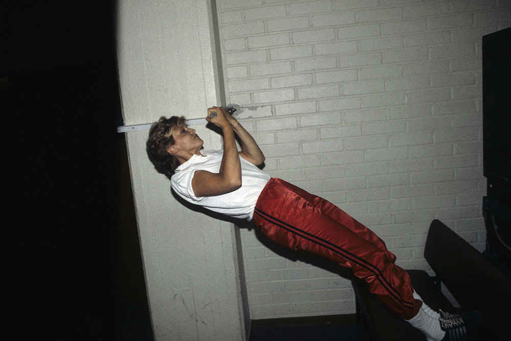 Färgbild av en kvinna i skjorta och sportbyxor som drar sig upp för ett hävräcke med fötterna på en bänk, inomhus. Tegelvägg i bakgrunden.