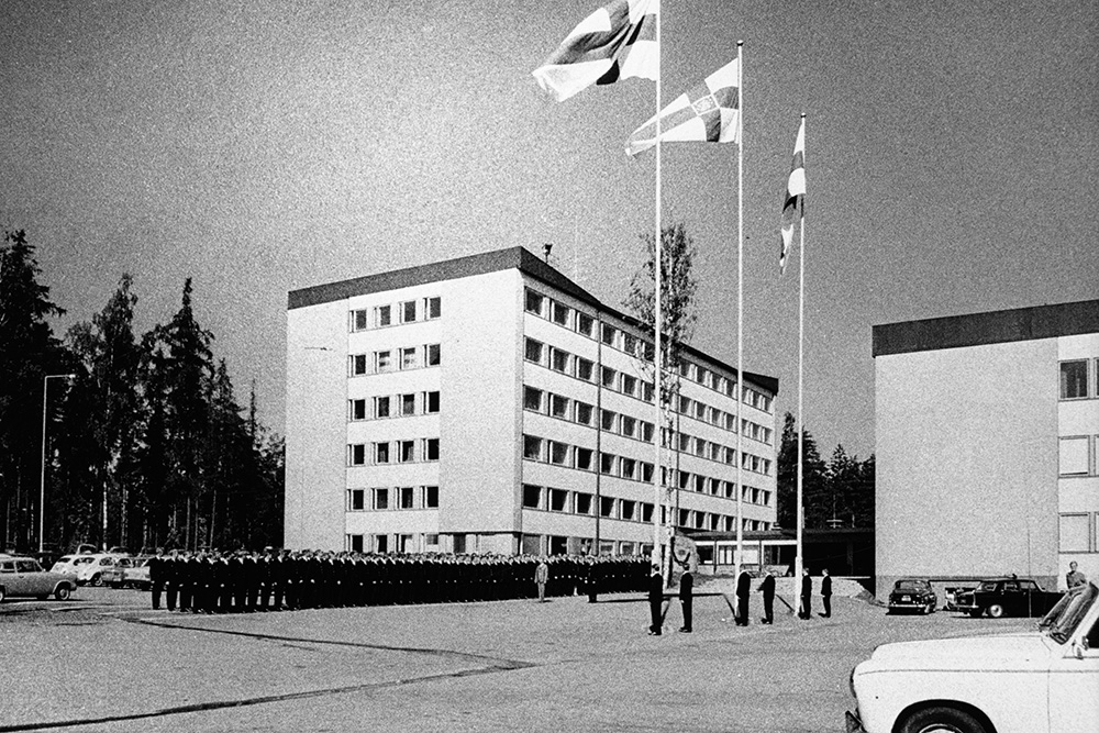 Dussintals uniformerade poliser står i rad för att se flaggan hissas på tre flaggstänger. Det svartvita fotot togs på en parkeringsplats, och bilden visar även flerbostadshus, träd och bilar.