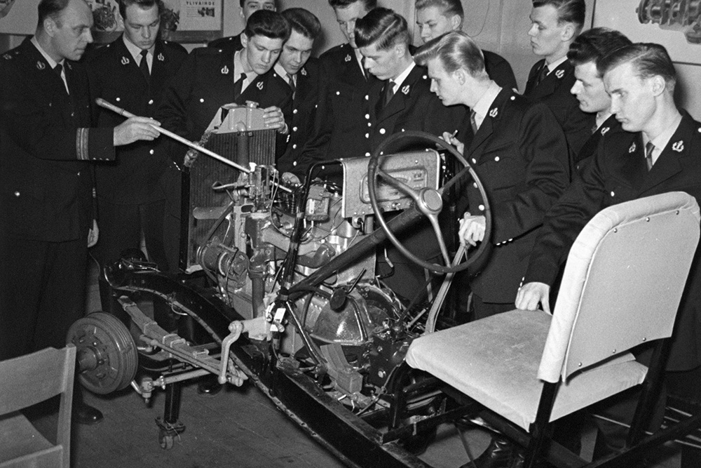 Ett svartvitt foto visar personer klädda i polisuniformer som undersöker delar av en bilmotor från en öppnad bilkaross. Till vänster pekar en person på motorn med en kartstav. Fotot togs i ett klassrum.