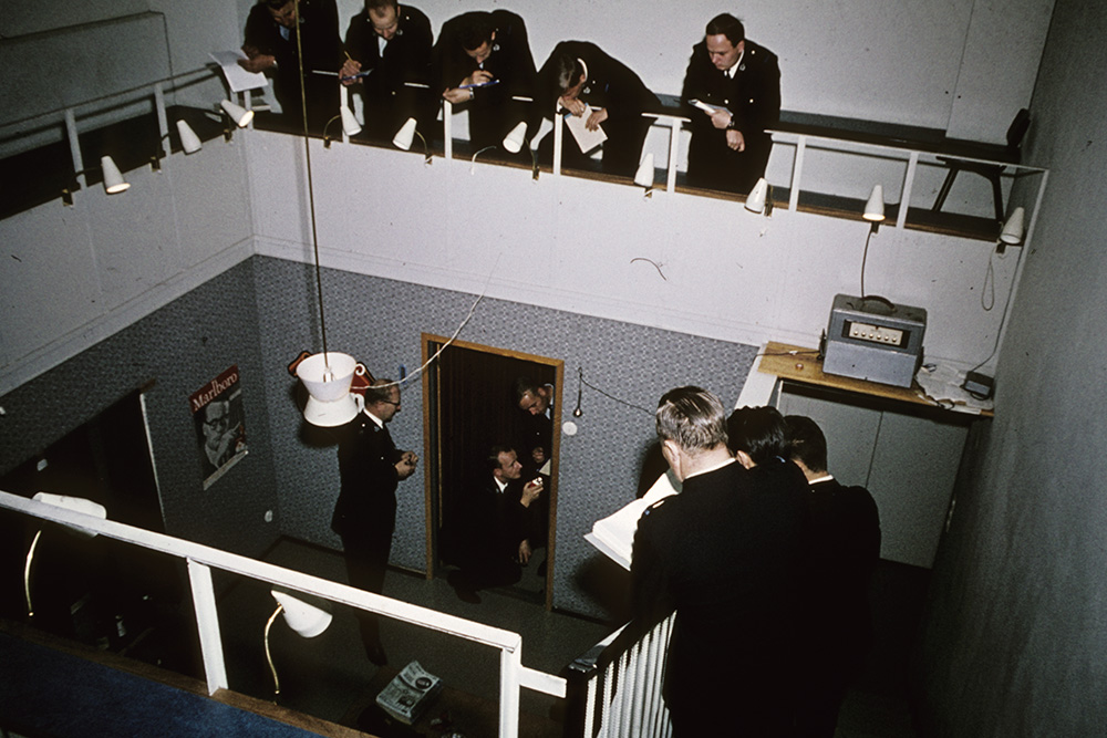 En färgbild av ett tvåvånings klassrum där personer klädda i polisuniformer tittar ner på en övningssituation från den övre läktaren, där tre personer undersöker dörrkarmarna. Klassrummet föreställer en lägenhet.