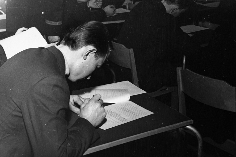 På den svartvita bilden skriver en man med en penna på vitt papper vid ett bord. I bakgrunden syns ett klassrum, med andra människor som sitter och är försjunkna i skrivande.