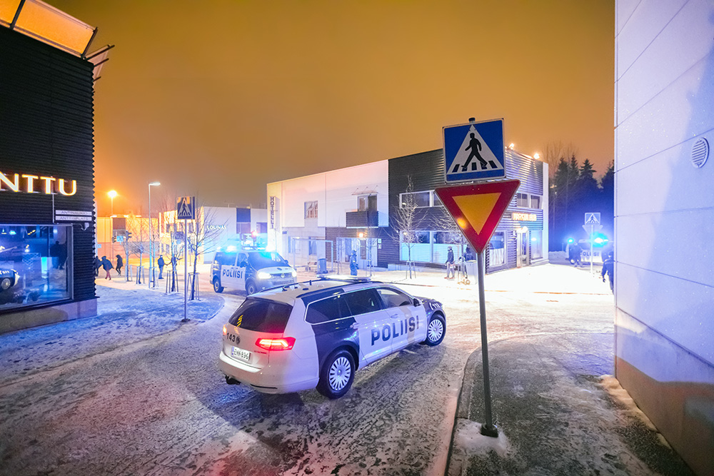Färgbilden visar byggnader, två polisbilar, fotgängare, trafikskyltar och träd i en stadsmiljö. Det ligger snö på marken.