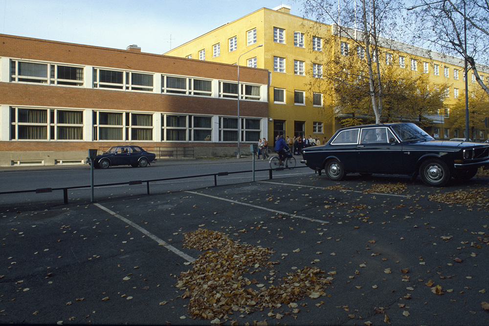 En färgbild av en parkeringsplats med en bil, träd och torkade löv på marken. Bakom finns en väg och två byggnader. Framför byggnaderna finns fotgängare, en cyklist och en bil.