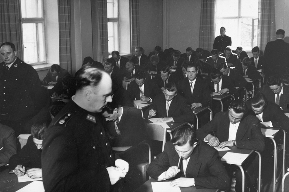 Den svartvita bilden visar dussintals människor som sitter och skriver på vitt papper i ett klassrum. Dessutom står fyra personer klädda i polisuniformer och övervakar händelsen. Två av personerna tittar direkt in i kameran.