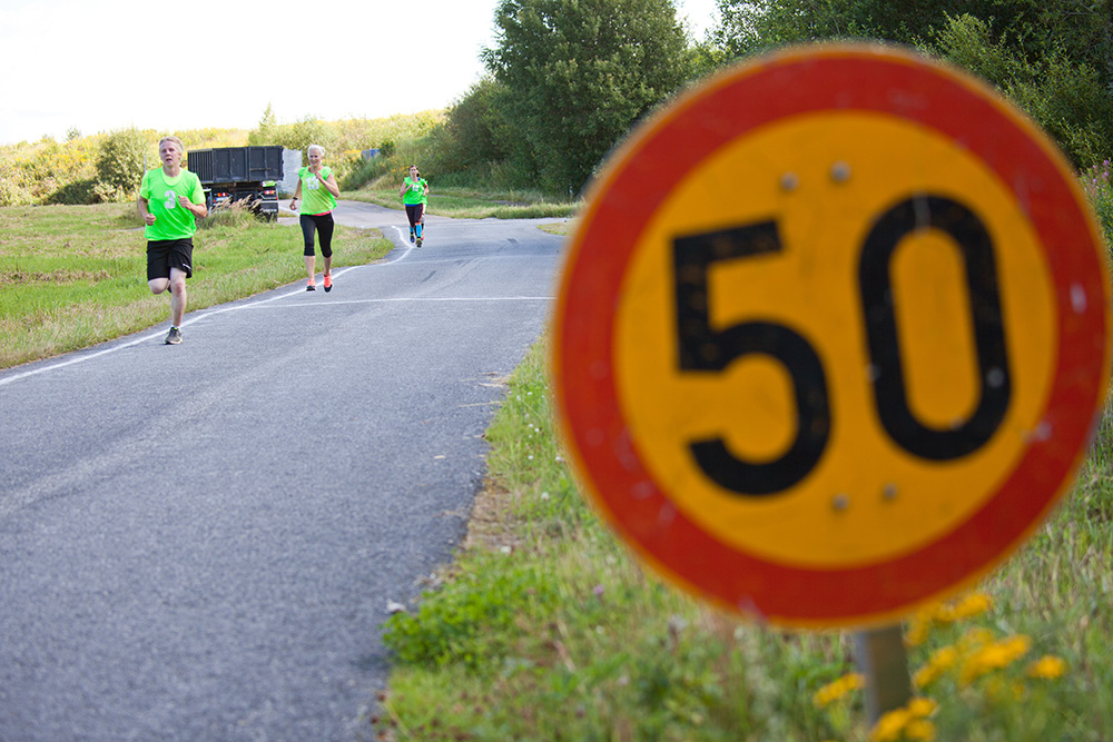 Färgbild av en asfaltväg. I förgrunden, till höger, finns en hastighetsbegränsningsskylt på femtio kilometer i timmen. Det finns löpare på vägen och i bakgrunden en lastbil, en äng och en skog. Löparna bär numrerade västar.