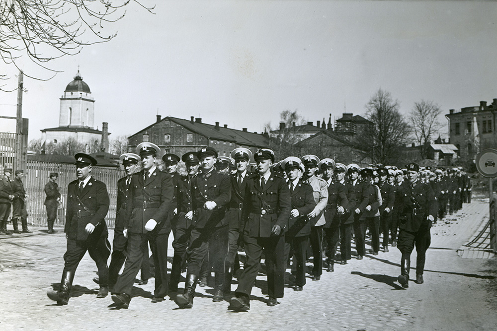 Den svartvita bilden visar människor klädda i polisuniformer som marscherar i formation. I bakgrunden syns en kyrka, andra byggnader, ett staket, en trafikskylt och träd. På vänster sida står tre personer klädda i militäruniformer.