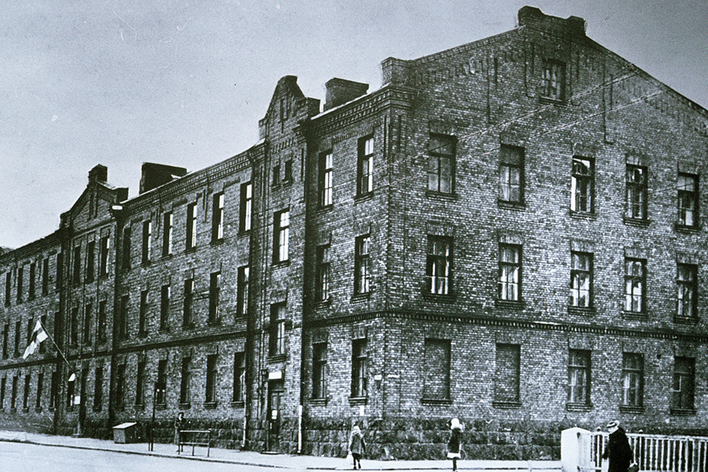 En svartvit bild av en trevånings stenbyggnad med fyra fotgängare som går framför den. Ovanför byggnadens ingång finns en korsflagga på en flaggstång.