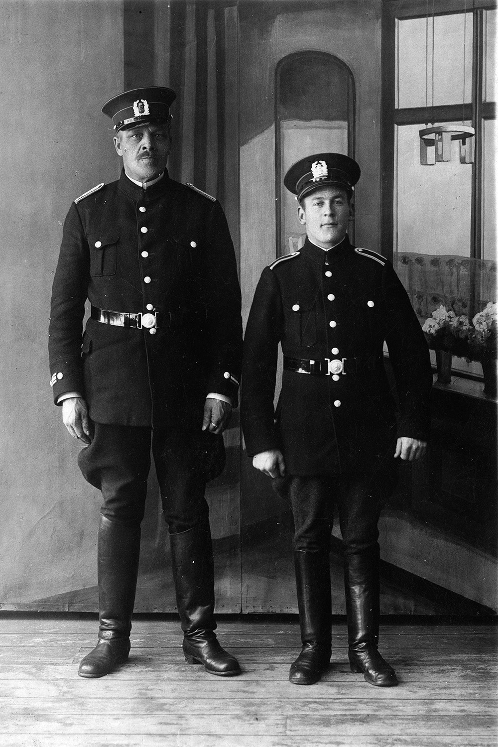 På det svartvita fotot poserar två personer i uniform vända mot kameran. I bakgrunden finns en vägg med teckningar som föreställer en fönstervy, gardiner och en fönsterbräda med två blomkrukor och en lampa på.