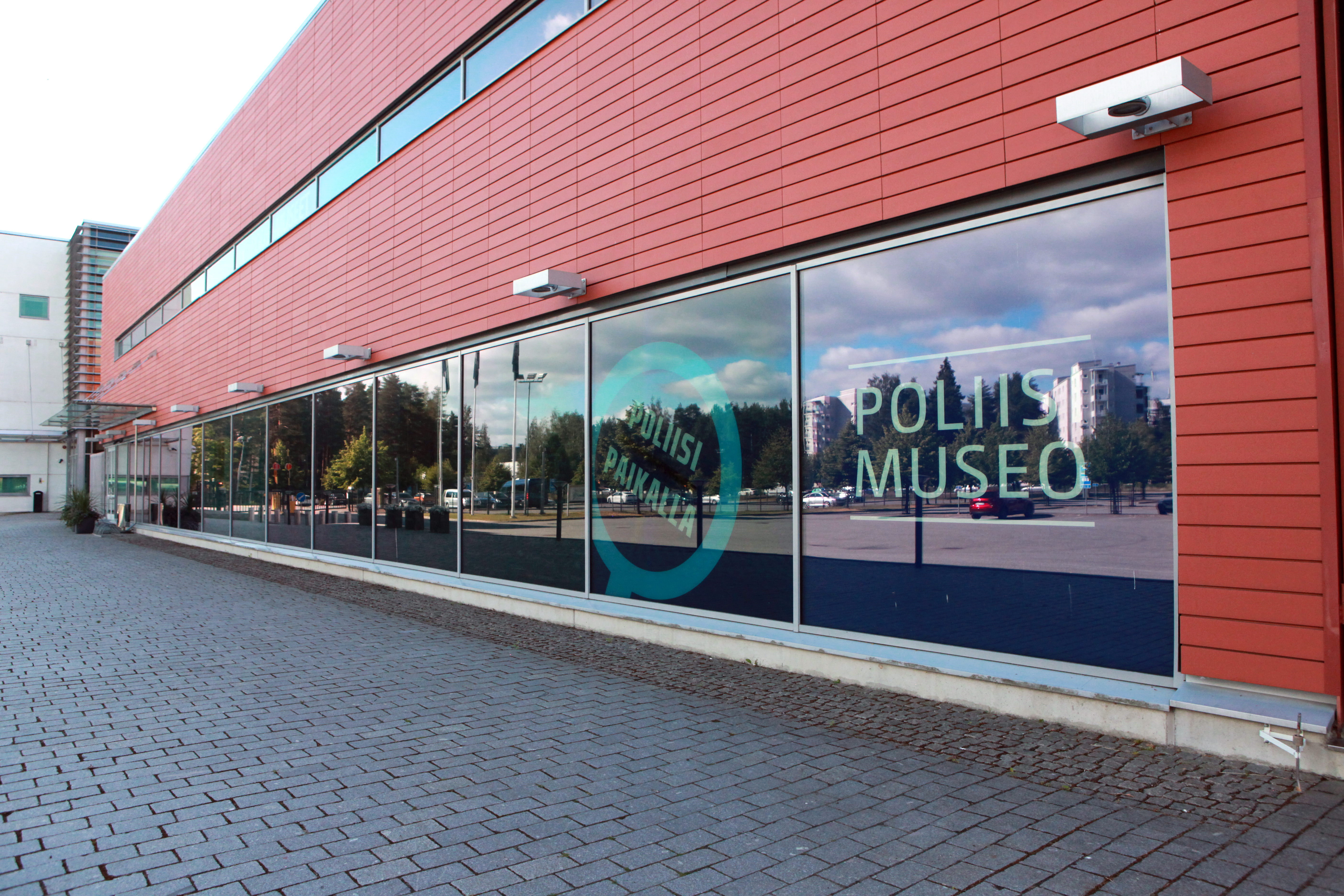 The front of the Police Museum building. The text “Police Museum” and the logo of the exhibition “The Police is Here!” are in the window, and the window reflects the parking lot opposite and houses. Photo The Police Museum, Pia Penttilä.