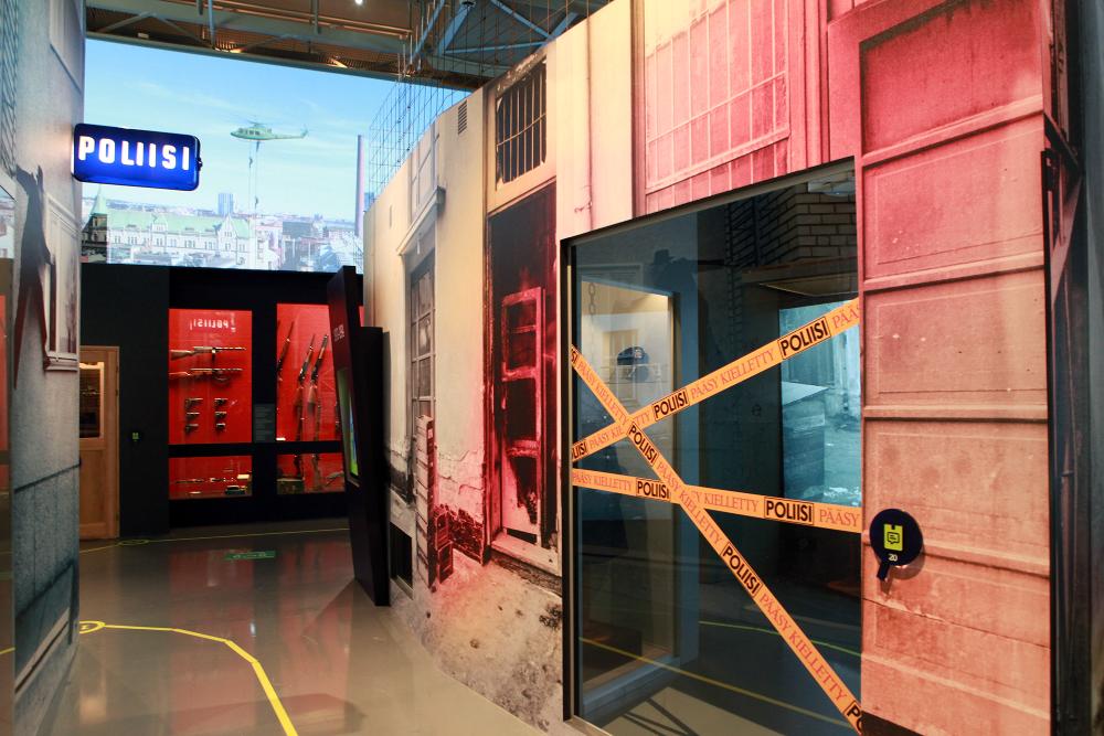 A view of the Museum’s exhibition. The picture shows, among others, the ‘Poliisi’ (the police) sign on the wall and some police tape “Police – do not cross”, and weapons on the wall in the background.