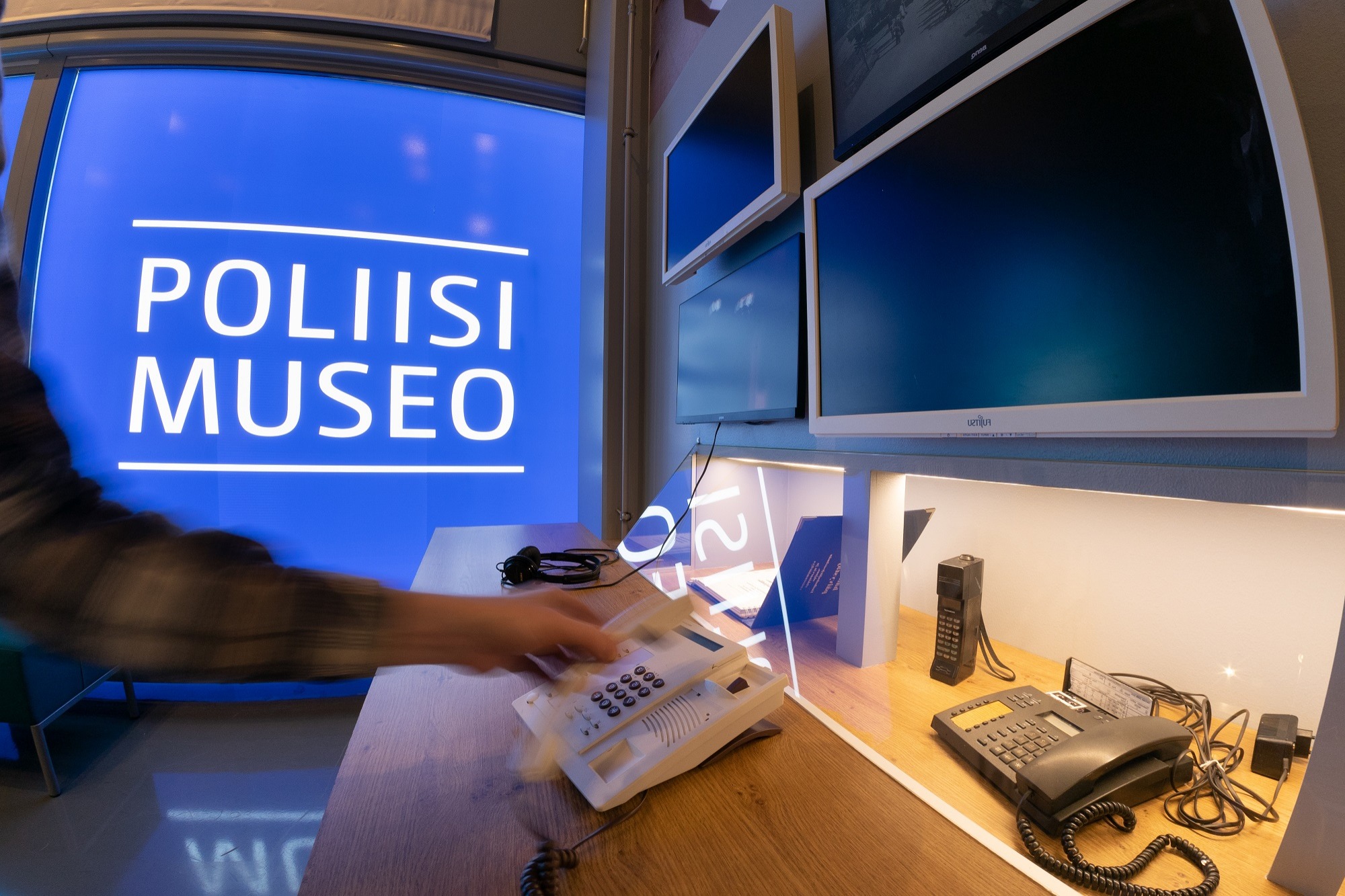 På bilden syns en hand som lyfter en telefon från bordet. I bakgrunden finns texten ”Poliisimuseo” (Polismuseet). Bild Polismuseet, Jarkko Järvinen