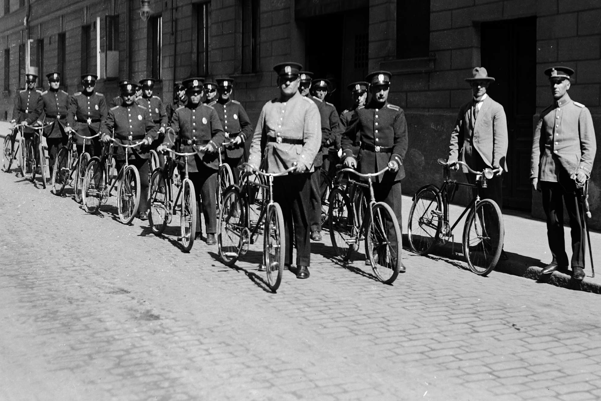 Mustavalkoinen vanha valokuva, jossa on virkapukuisia poliiseja polkupyörineen. Poliisit seisovat kahdessa jonossa kaupungin kadulla. Kuva Poliisimuseo