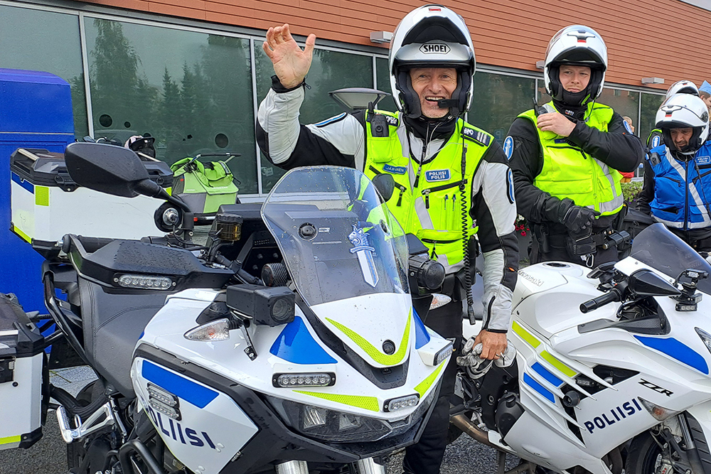 A motorcycle patrol officer in riding gear waves to the camera. Next to the waving police officer, there are police motorcycles and other police officers.