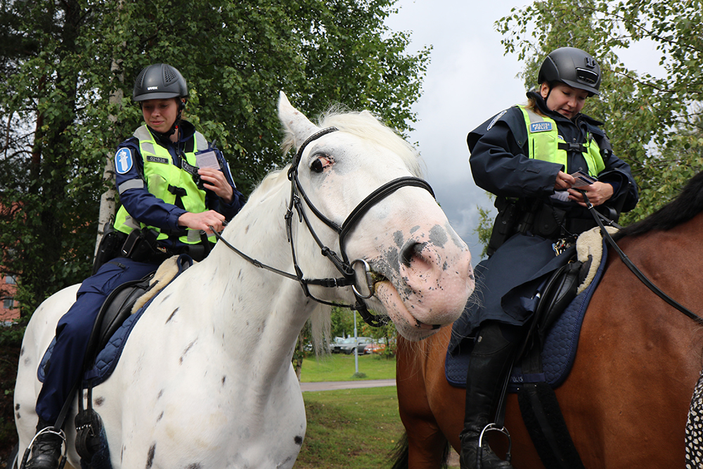 Kaksi ratsupoliisia istuu poliisihevosten selässä, taustalla puita ja piha-aluetta.