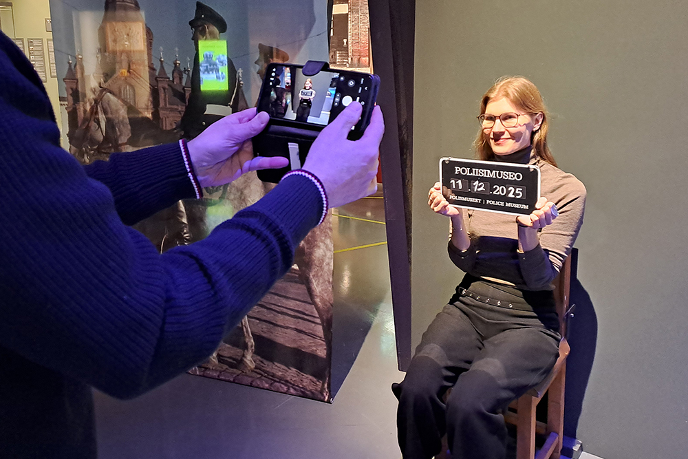 Kameraan selin oleva henkilö ottaa valokuvaa toisesta henkilöstä, joka istuu puisella, vanhalla tuolilla. Istuvalla henkilöllä on kyltti, jossa lukee "Poliisimuseo" ja päivämäärä. Taustalla näkyy museon näyttelyä.
