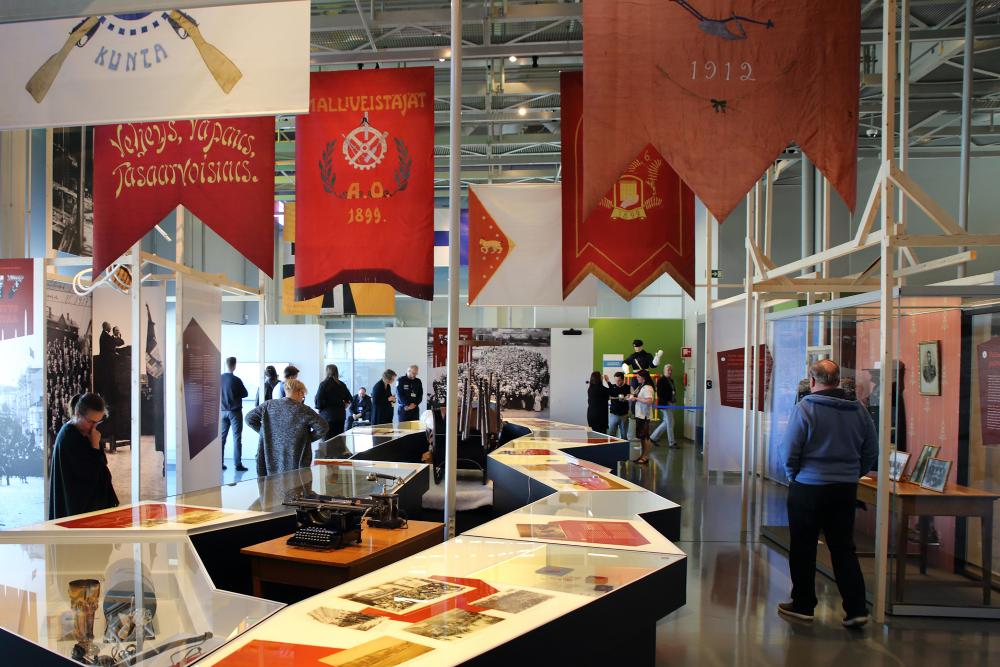 General view of the exhibition “Public order collapses 1917”. The picture shows museum visitors viewing the exhibition, with old banners and photographs in the foreground.