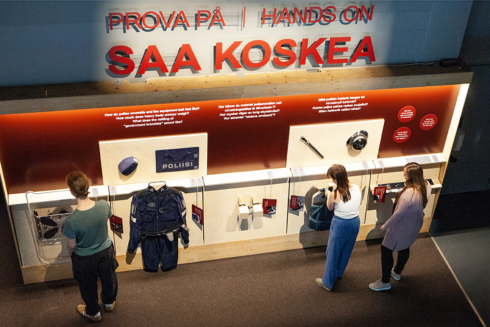 Three people by the Police Museum’s wall of items that may be touched. The police tools and equipment on the wall may be examined and touched.