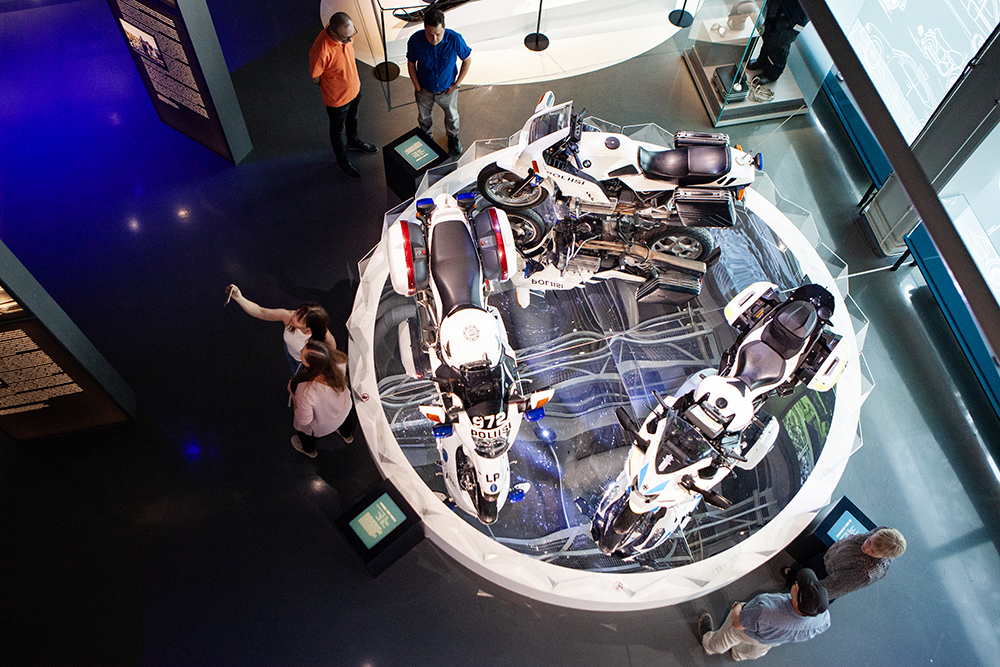 Six people around the motorcycles on display in the museum’s exhibition space. The image is taken from above. The motorcycles are on display on a podium with mirrors on the floor that reflect the ceiling structures.