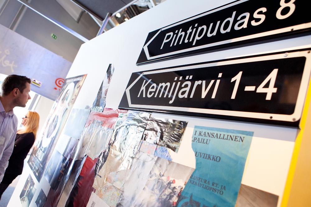 Two museum visitors are viewing the exhibition. In front in the picture, there are signs to various towns.