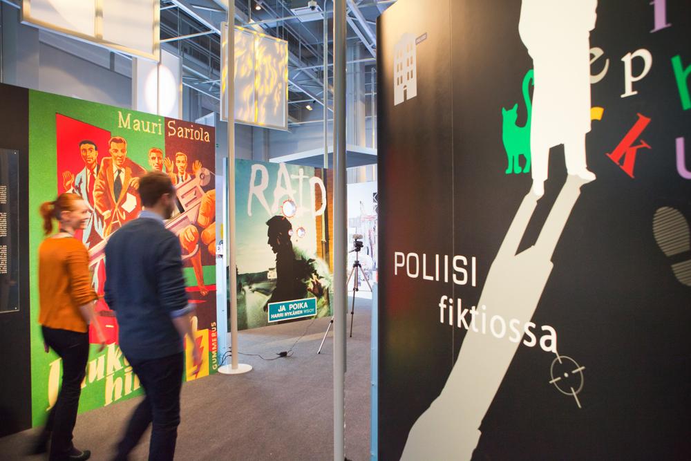 Yleiskuva Poliisi fiktiossa -näyttelystä. Kuvassa kaksi museovierailijaa kävelemässä näyttelyyn, ympärillä näyttelyyn liittyviä piirroskuvia.