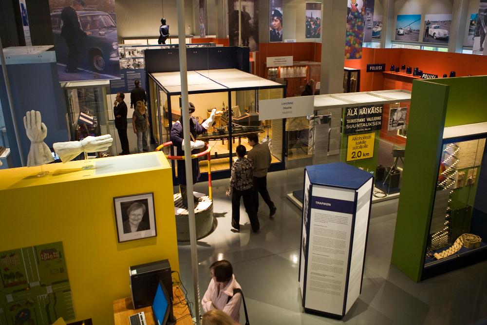 General view of the exhibition “900 years of police history in Finland”, in a photo taken near the ceiling. The picture shows museum visitors walking between the showcases, and in the room, for example photographs and items relating to police work are seen.